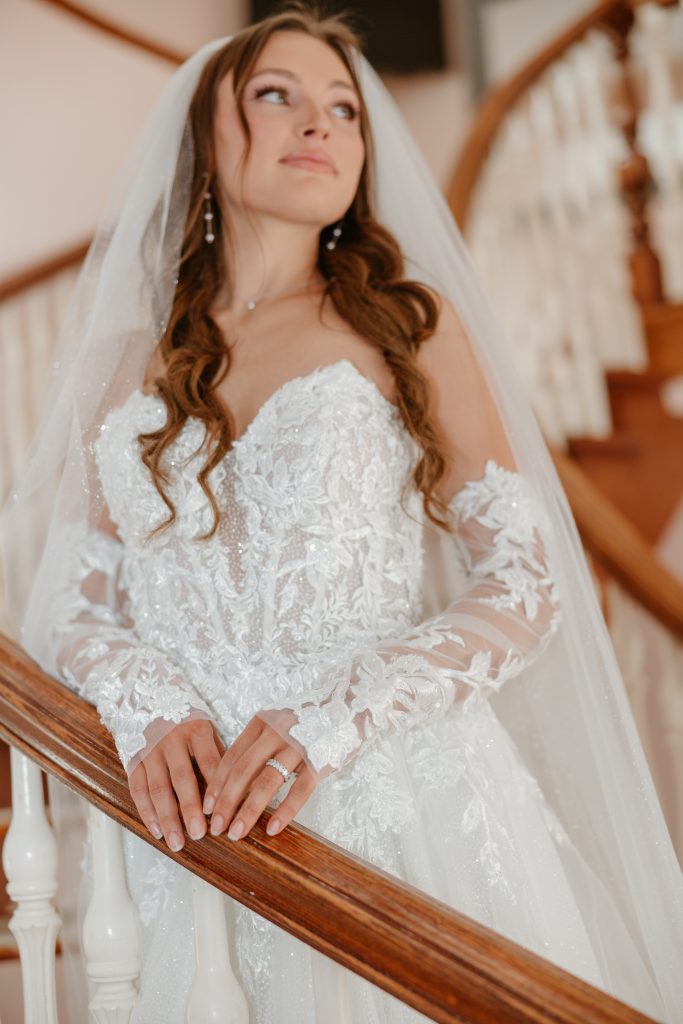 A bride in a lace wedding gown, sheer floral sleeves, and a veil poses beside a curved wooden staircase.