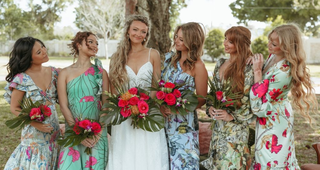 A bride in a white wedding dress stands with five women in colorful floral dresses, each holding a vibrant tropical bouquet.