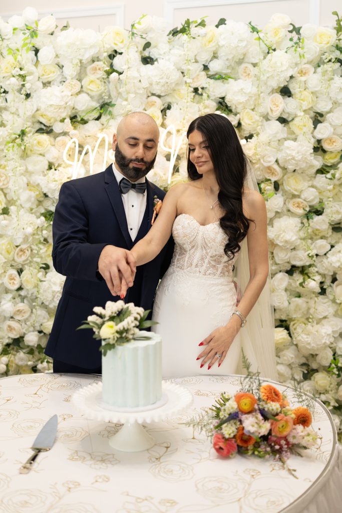 Bride and groom cutting their wedding cake in front of a wall filled with white flowers.