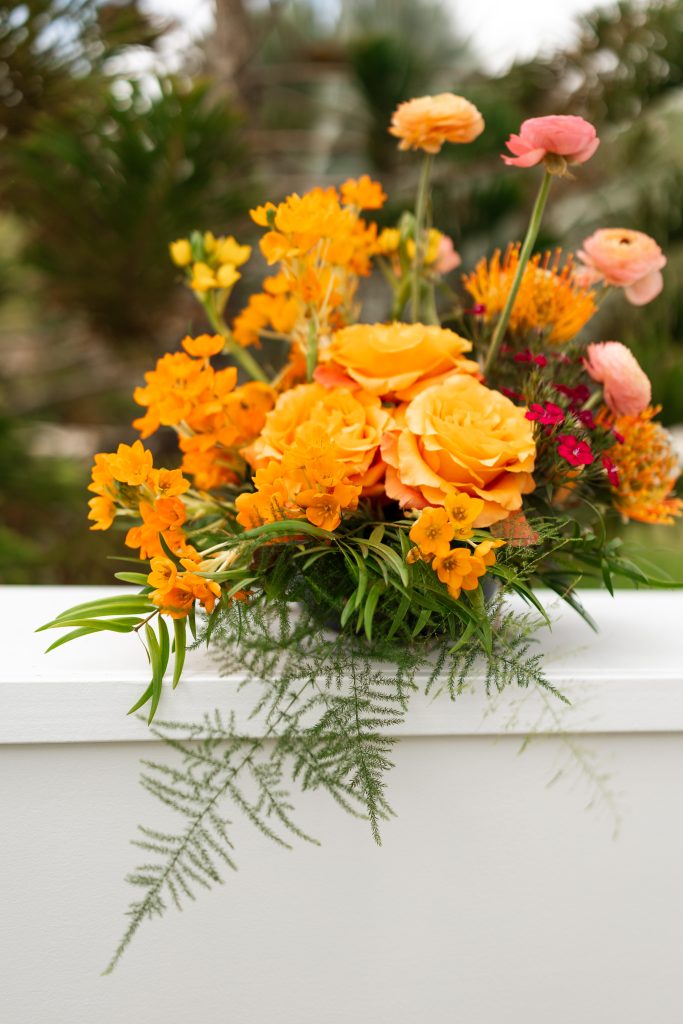Orange and peach floral arrangement with roses, greenery, and colorful accent flowers.