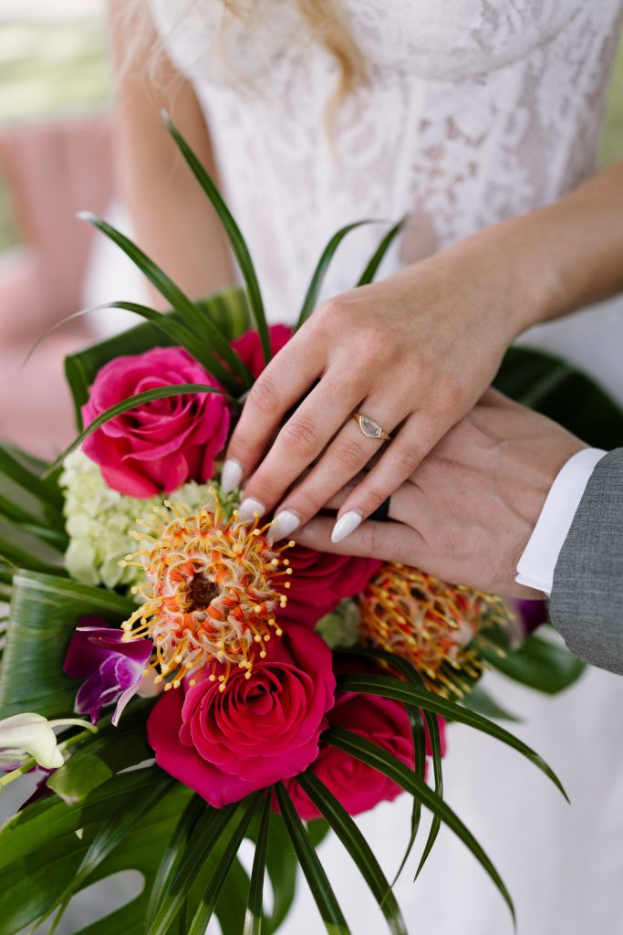 A close-up shows a couple’s hands resting over a bouquet of bright pink roses, orange flowers, and tropical greenery.
