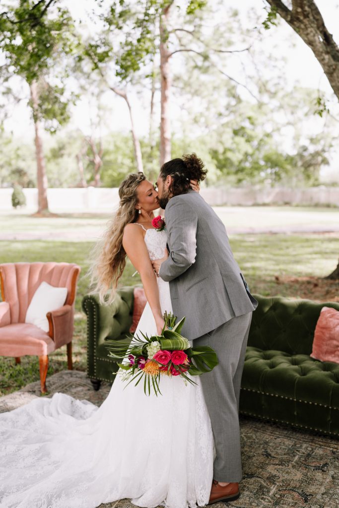 A bride in a lace wedding gown holds a tropical bouquet while kissing a groom in a gray suit beside outdoor lounge furniture.
