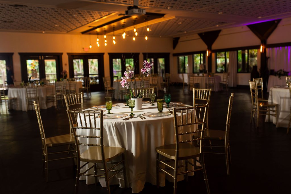 A reception table with gold chairs and an orchid centerpiece sits in a warmly lit event space.