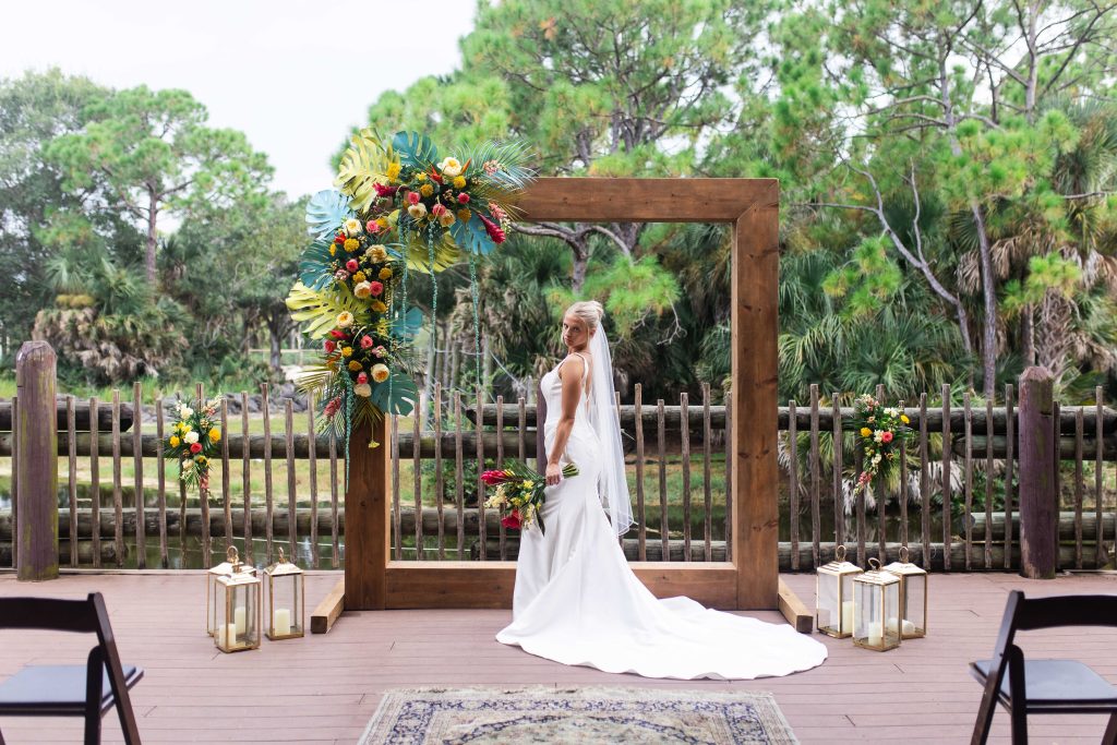A bride poses beneath a wooden ceremony frame decorated with colorful tropical flowers.