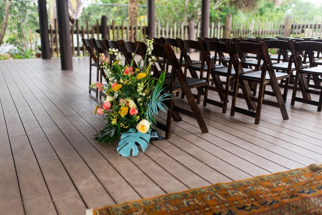 A colorful floral arrangement sits beside rows of dark wooden chairs on an outdoor deck.