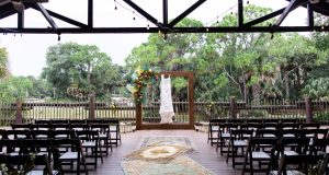Rows of dark wooden chairs face a floral ceremony frame with a wedding dress hanging from it.