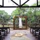Rows of dark wooden chairs face a floral ceremony frame with a wedding dress hanging from it.