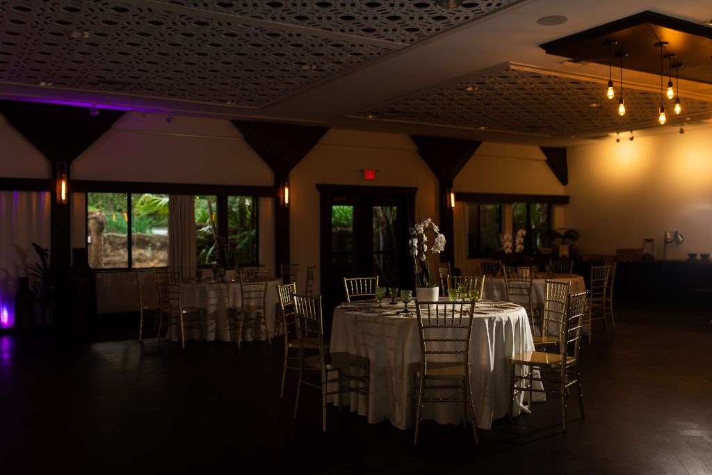 Round reception tables with gold chairs fill a dimly lit event room with warm pendant lights.