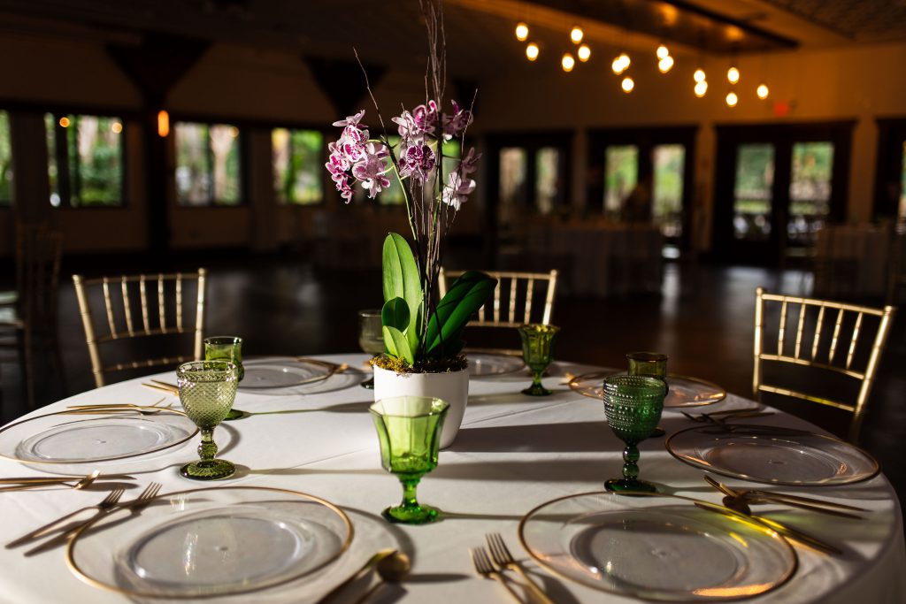 An orchid centerpiece, green glasses, clear plates, and gold flatware decorate a reception table.