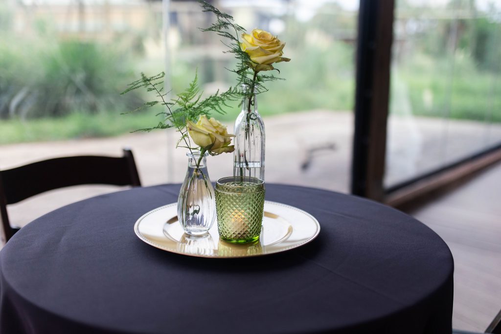 Two yellow roses in glass vases and a green candle holder decorate a black-covered table.