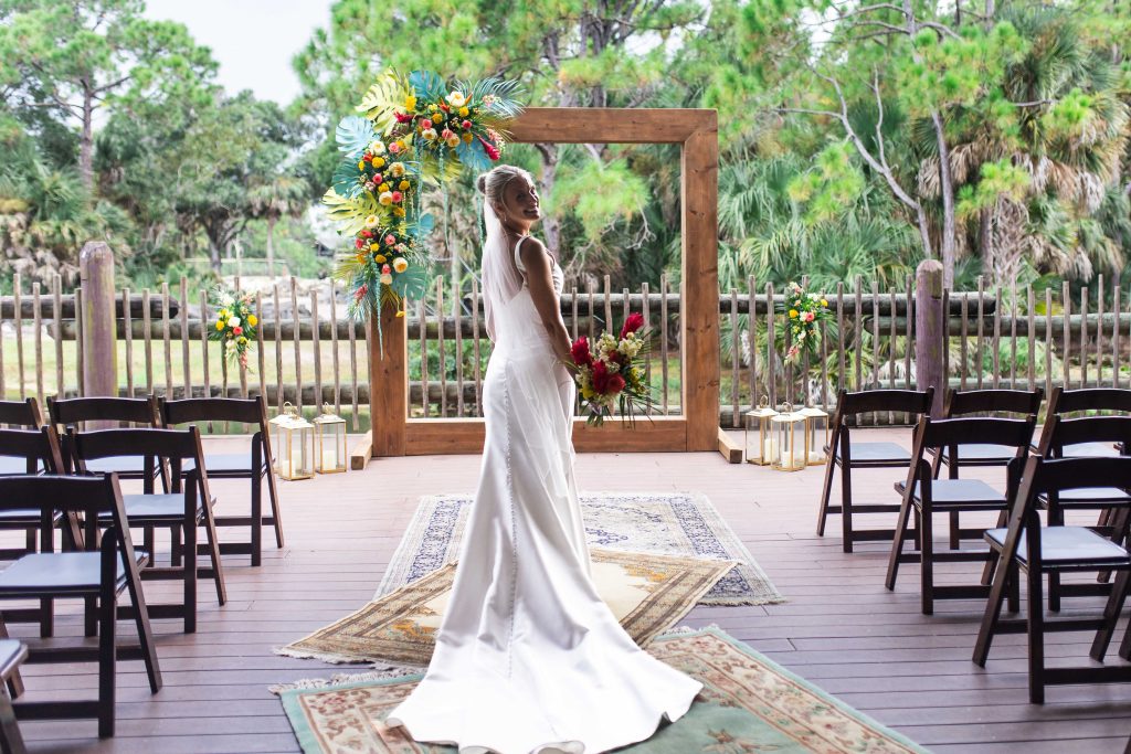 A smiling bride holding a bouquet stands beneath a floral wooden ceremony frame.