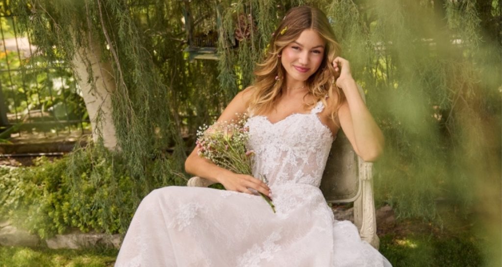 A woman sits outdoors in a garden wearing a lace wedding dress while holding a small bouquet of pink and white flowers.