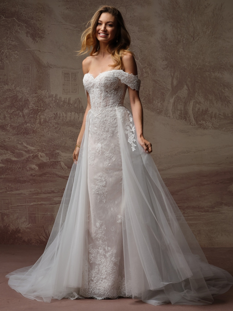 A smiling woman wears an off-the-shoulder lace wedding dress with a fitted silhouette and a flowing sheer tulle overskirt.