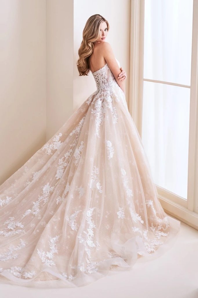 A woman poses by a bright window in a strapless wedding gown with a corset-style back, floral appliqu&eacute;s, and a long tulle train.