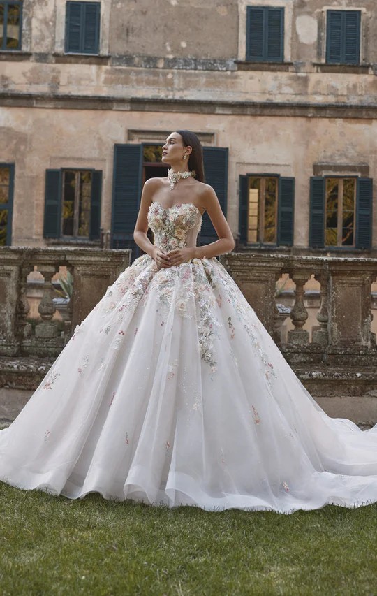 A woman wears a strapless ball gown with colorful floral appliqu&eacute;s and a voluminous tulle skirt outside a historic stone building.
