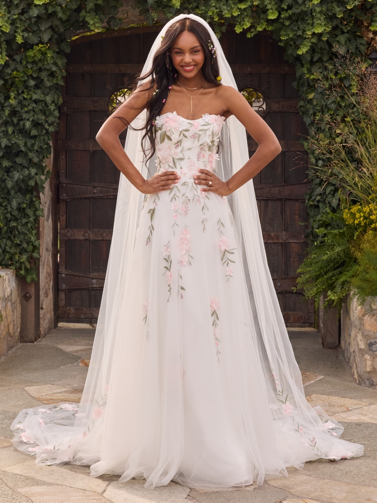 A woman poses in front of ivy-covered wooden doors wearing a strapless wedding dress with pink floral embroidery and a long sheer veil.