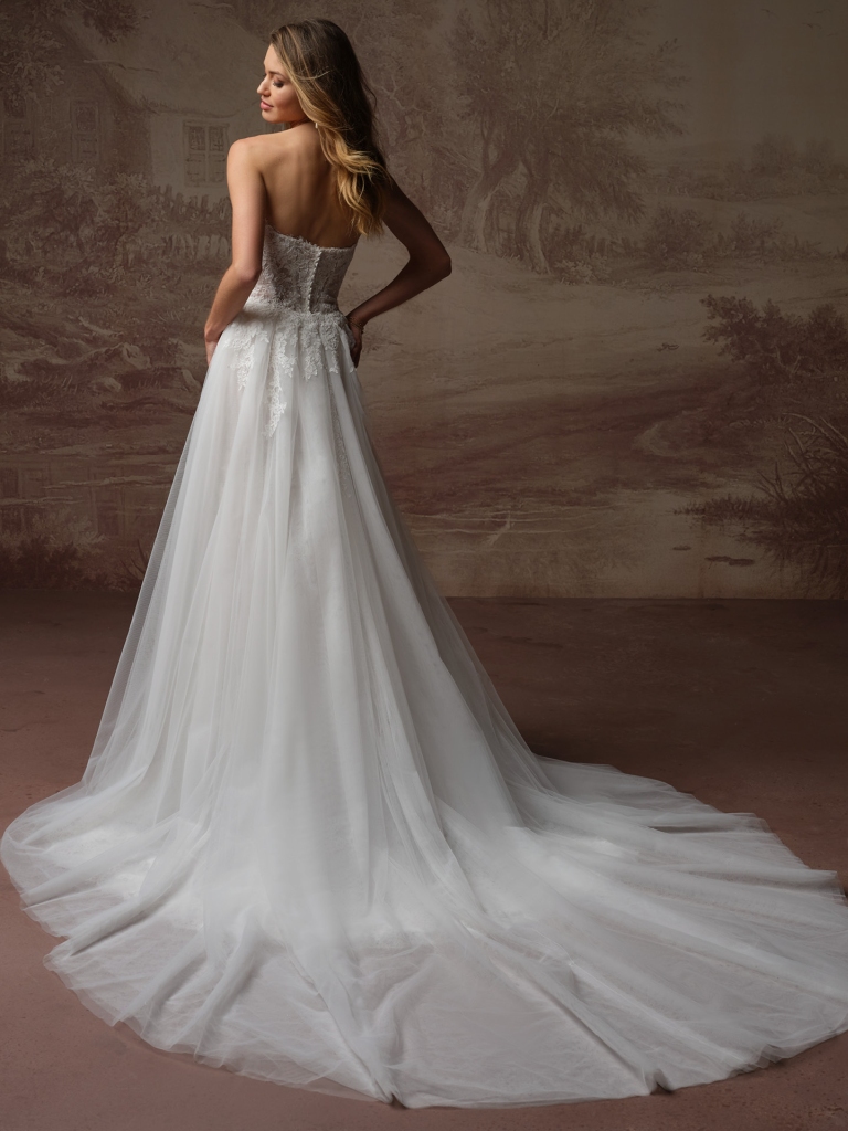 A woman is shown from behind wearing a strapless wedding gown with pink floral embroidery and a flowing skirt against a neutral studio backdrop.