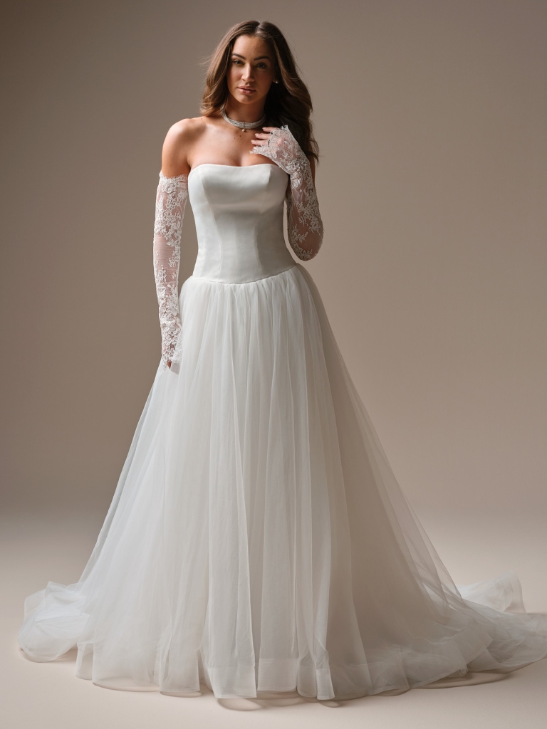 A woman models a strapless wedding gown with a structured satin bodice, full tulle skirt, and long lace gloves against a neutral studio backdrop.